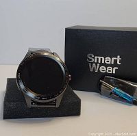 Black metal Smart Wear branded smartwatch displayed on black foam stand next to its charging cable and box