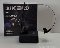 Photo of Aikela BH-M9A mono wireless headset positioned on charging base with the product box behind it, showing the brand and model information.