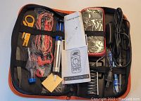 Zippered orange and black carrying case opened to display multiple car safety and repair tools including soldering iron, digital multimeter manual, wires, cables, and hand tools all organized with elastic straps inside.