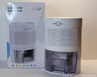White Lonove dehumidifier beside its original packaging box. Device features a transparent water tank and a simple rotary control knob on the front.