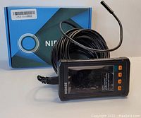 Photo shows a Nidage dual lens endoscope with a long black cable, a small camera tip at the end, and a monitor unit. The monitor has an LCD screen and four orange buttons.