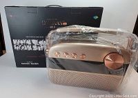 Photo of Saregama Carvaan 2.0 Gold with original retail box behind it. Device is wrapped in protective plastic on top with visible gold knobs and handle.