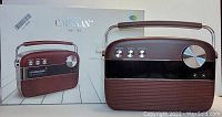 Saregama Carvaan Hindi radio with brown housing, control knobs for Anthems, Modes, and Day/Night, and large volume knob. The radio is standing next to its original box with product image and branding.