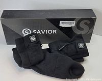 Photo shows a pair of black rechargeable heated socks in front of their black Savior branded box.