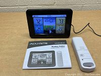 Acu-Rite weather station with color digital display, outdoor wireless sensor, and instruction manual placed on wooden table against white brick wall.