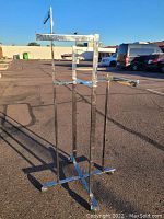 Metal clothing rack shown standing on asphalt with height adjustable vertical bars and horizontal bars for hanging clothes, no clothes present.