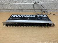 Front view of Behringer Multicom Pro MDX 4400 showing control knobs and power cable on wooden surface against brick background.