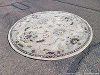 Photo of large round area rug on outdoor ground, showing floral and leaf pattern in neutral tones with decorative border.