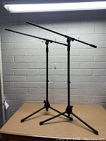 Two assembled black On Stage boom microphone stands side by side on a wooden table against a white brick wall, showing the tripod bases, adjustable vertical poles, and horizontal boom arms.