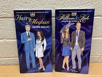 Full photo showing both Harry & Meghan and William & Kate paper doll books standing upright against a beige wall, covers visible.