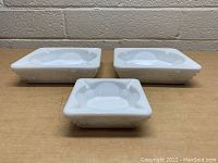 Front and side perspective of all three square milk glass ashtrays showing floral detail and shape.