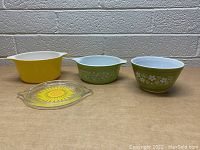 Three Pyrex dishes and one glass lid displayed from front, showing daisy floral patterns and yellow dish with sunflower lid