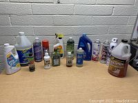 Photo shows a group of 11 various cleaning bottles including spray, pump, and aerosol cans on a wooden table against a white brick wall.