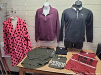 Six assorted Victoria's Secret Pink clothing items displayed on and around two mannequins and a wooden table against a white brick wall