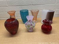Front view of seven colorful glass vases arranged on a plain surface against a light brick wall.