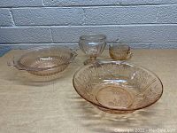 Four pink glass pieces arranged on a table: large patterned bowl, footed bowl, ribbed bowl with handles, and a small sugar bowl with handle.