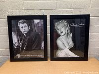 Front view of two black-framed posters under glass featuring James Dean and Marilyn Monroe placed side by side on a wooden surface against a brick wall.