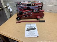Red hydraulic floor jack with black wheels and lifting pad, curved detachable handle, instruction manual, and original box on a table.
