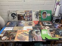 Photo showing 19 vinyl records laid out on a table with visible colorful album covers featuring artists in country, jazz, blues, pop genres.