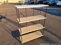 Four-tier shelf unit with four flat shelves in light wood or laminate finish, supported by a black metal frame with diagonal and vertical supports. The unit is photographed outdoors in natural light from two angles, showing its structure and proportions.