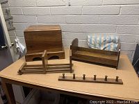 Photo showing all four wooden items on a wooden table against a brick wall background: small chest, napkin holder, bookend/magazine holder, and rack with knobs.