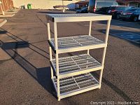 Front angle view of the plastic storage shelf unit outside on pavement with cars in background, showing four grated shelves and tubular legs.