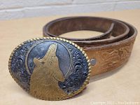 Leather belt with embossed western horse and rider design next to oval belt buckle on wooden surface