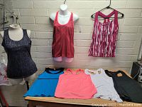 Six women's activewear tops in different colors and styles laid out and on mannequins against a brick wall, all size large.