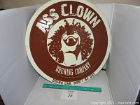 Front of round metal beer sign with Ass Clown Brewing Company logo and slogan.