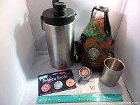Metal growler with black lid beside glass growler in neoprene jacket, metal cup, bicycle bell, and pin set.