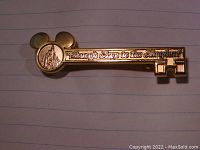 Close-up photo of the gold-colored metal key-shaped pin engraved with 'Disney's Keys to the Kingdom' and Mickey ears on top
