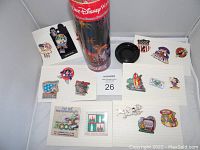 Overview of laid out Disney cast member pins displayed on white lined cards arranged around a red cylindrical container for a Disney Coca-Cola commemorative bottle. Pins include limited edition, patriotic themed, and character pins.