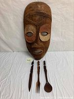 Wooden tribal mask and three carved wooden utensils (fork, serving spoon, cutting utensil) laid out on a white cloth.