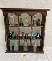 Wooden wall hanging shelf holding 14 various collectible glass bottles arranged in compartments.