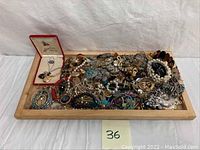Photo shows large assortment of costume jewelry piled on a wooden tray including brooches, chains, earrings, pearl-like bead necklace, and tortoiseshell-effect accessories.