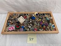 Top-down view of a wooden tray filled with assorted costume jewelry pieces including brooches, pins, earrings, and pendants in multiple colors and materials