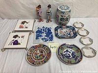 Full lot display of Oriental porcelain and ceramics items including covered jar, Geisha figurines, plates, trays, salt dishes, and tile trivets on a white backdrop.