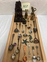 Overview of all items displayed on wooden tray including souvenir spoons, brass cat, brass bell, bronze elephant, resin figurines.