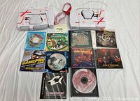 Two Lazer X laser tag shooters and 9 video game discs laid out on white surface. Shooters are white with grey grips and orange/red details, with wrist straps attached. Video game discs include various titles visible in top view.