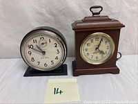 Two vintage tabletop clocks side by side on a white cloth background; left is the Westclox Big Ben in black metal case, right is the Seth Thomas wooden mantel clock.