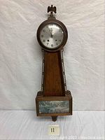 Full view of vintage Gilbert banjo wall clock standing on white backdrop. Shows overall shape and painted scenic lower panel.