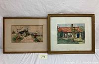 Two framed watercolor paintings side by side on a white backdrop. Left painting depicts a country road and mailbox scene, right painting shows a house with red roof and blue doors.