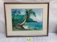 Framed watercolor painting showing tropical beach scene with palm trees, ocean, and mountains in background.