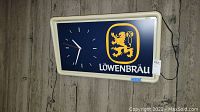 Front view of Löwenbräu clock showing white frame, blue face, clock hands and attached power cord on wooden background.