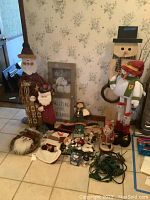 Wide view of the lot showing hand painted wooden Santa, large snowman welcome sign, assorted figurines, and miscellaneous holiday decorations arranged on floor.
