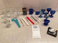 Photo showing a collection of blue and clear glass eyewash cups, four glass thermometers with colored tops, a vintage compact with floral decoration, and a blue plastic case, all arranged on a countertop.