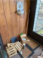 Wide view showing wall mounted fountain above, set of five wooden plant dollies, coir floor mats, cardboard box with various items, metallic cylinder container, hedgehog boot scraper.