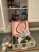 White cabinet shelf with various golf balls in boxes, cans, and loose along with a baseball cap, a vintage wooden tennis racket leaning against a white carrying case filled with assorted sports balls and golf items.