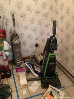 Overview of vacuum cleaners including Panasonic upright vacuum, Shark upright vacuum, Eureka handheld vacuum, attachments and vacuum bags on tiled floor against floral wallpapered wall.