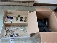 Overhead photo showing an open box filled with vintage eyeglasses and sunglasses with some frames displaying blue, green, black and brown lenses, and a separate box containing multiple modern black eyeglass cases.